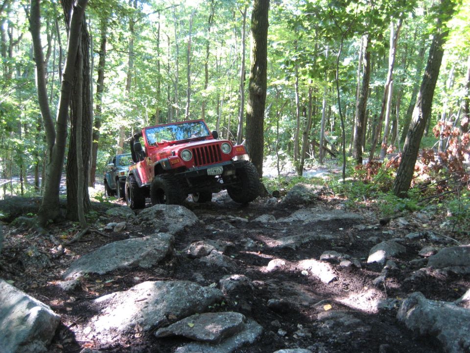 Red 2003 Jeep Wrangler navigating a rocky trail, front-left tire lifted