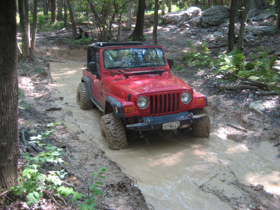 Red 2003 Jeep Wrangler driving through a muddy trail in a forest
