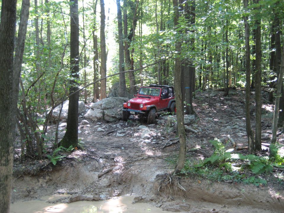 Red 2003 Jeep Wrangler navigating a rocky, muddy forest trail