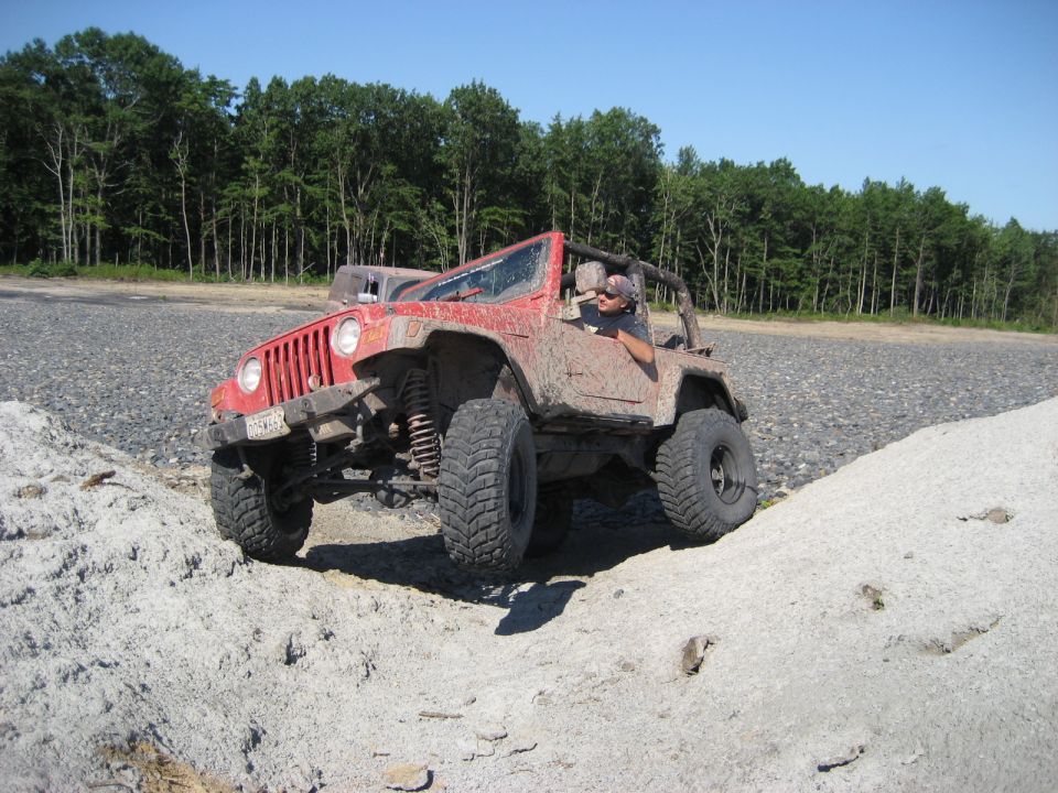 Red Jeep Wrangler driving over rough terrain with trees in the background. Driver is visible