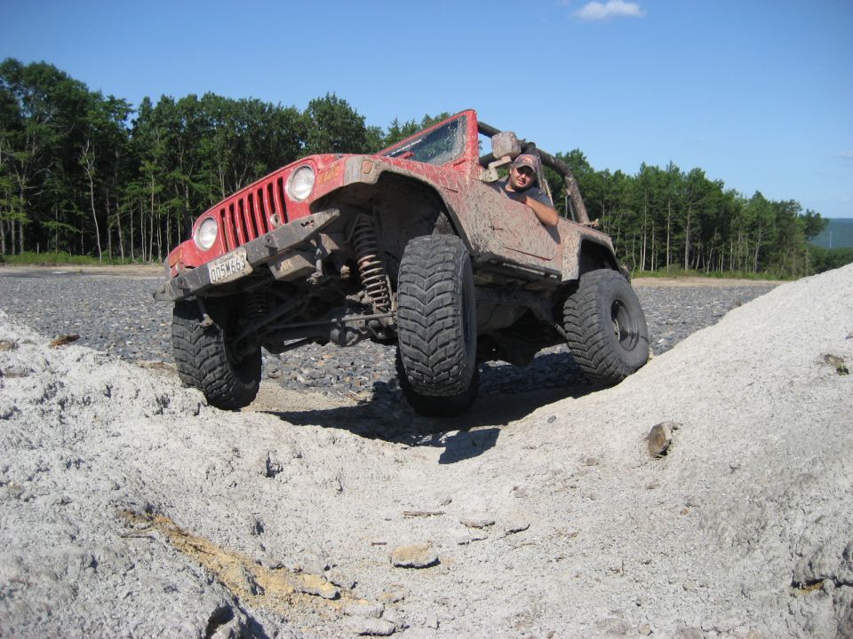 Red 2003 Jeep Wrangler with large tires climbing a dirt mound, driver visible