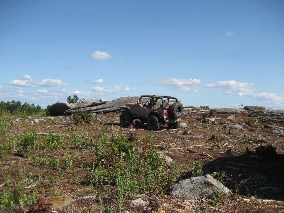 Red Jeep Wrangler parked on rocky terrain with trees and blue sky