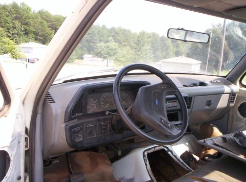 1995 Ford Bronco - Gutted interior of a Ford truck shot from the driver's doorway, showing the gray dash, four-spoke steering wheel with Ford emblem, analog g…
