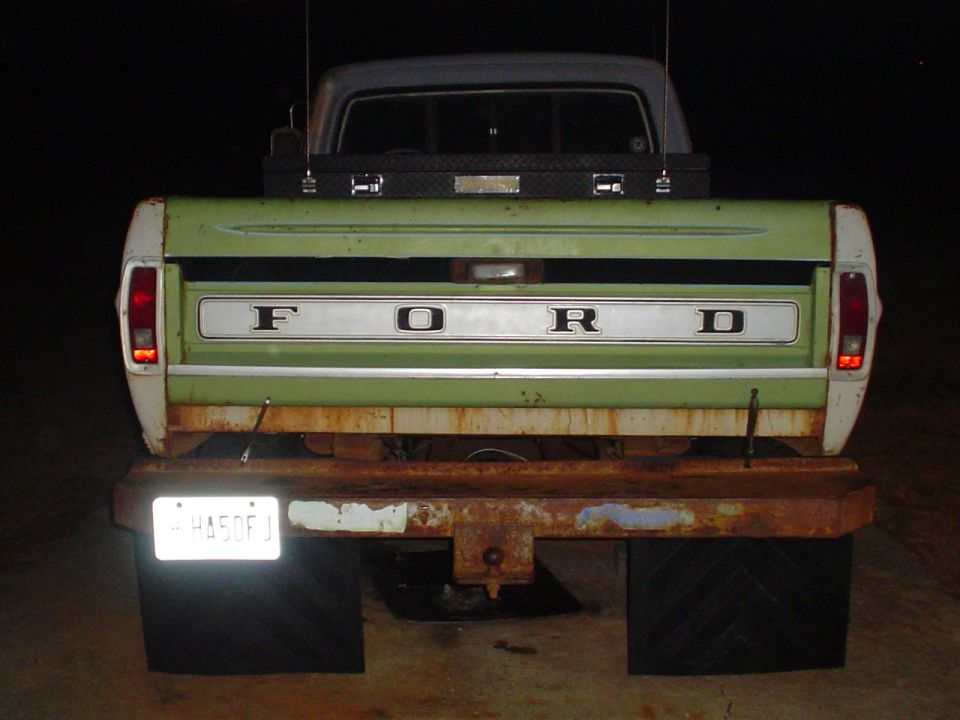 Green and white Ford pickup truck tailgate with chrome trim and "FORD" lettering. Rusty bumper and black mud flaps visible