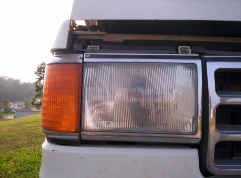 1995 Ford Bronco - Close-up of the passenger-side rectangular headlight and amber wraparound corner marker on a white Ford truck, hood raised to show the latc…