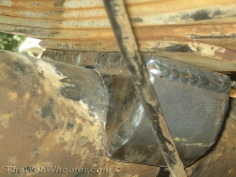 1990 Ford Bronco - Close-up of welded steel suspension fabrication on a dirty axle tube with leaf spring coils visible above.