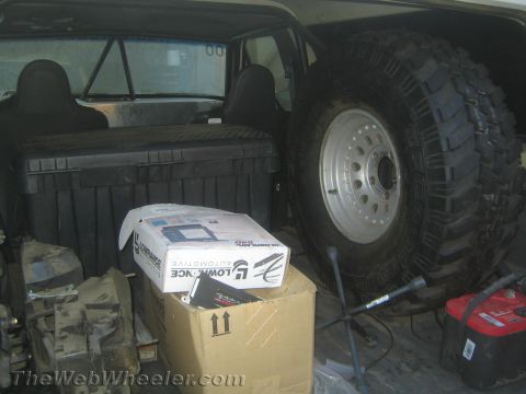 1990 Ford Bronco - large spare off-road tire on a white steel wheel and a parts box loaded in the cargo area behind a vehicle's seats.