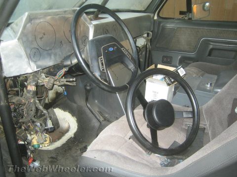 1990 Ford Bronco - gutted vehicle interior showing a bare fabricated metal dash, steering wheel, cloth seat and exposed wiring harness.
