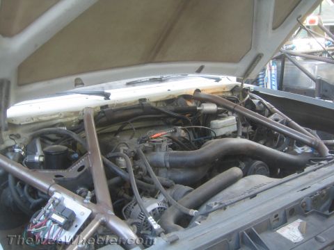 1990 Ford Bronco - Engine bay of a project vehicle showing tubular cage members, a V8 engine, exhaust manifolds and hoses under a raised hood.