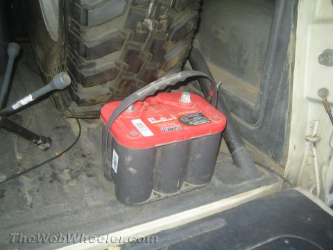 1990 Ford Bronco - red-top spiral-cell battery mounted on a dirty vehicle floor next to a chunky off-road tire and driveshaft.
