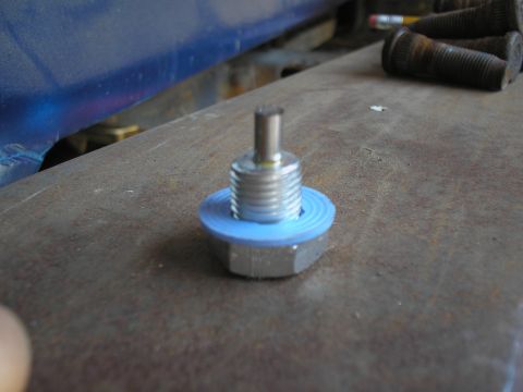 1990 Ford Bronco - Close-up of an oil drain plug with a blue gasket, resting on a textured metal surface.