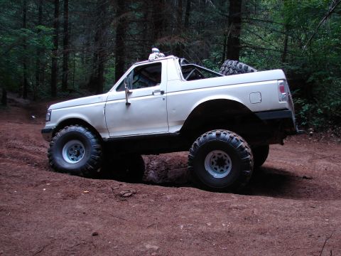 1996 Ford Bronco - White Ford Bronco with off-road modifications driving on a dirt path surrounded by trees.