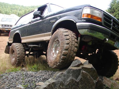 1996 Ford Bronco - A lifted dark blue 1996 Ford Bronco navigates rocky terrain, with another white vehicle visible in the background.