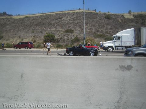 1990 Ford Bronco - roadside crash scene on a divided highway with a damaged black convertible, debris, a semi-truck and people gathered.