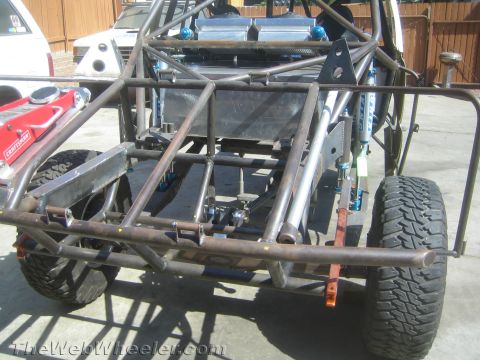 1990 Ford Bronco - Rear view of a bare tube-chassis off-road race buggy with blue coilover shocks and skid plate, parked in a driveway.