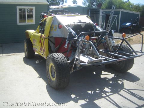 1990 Ford Bronco - yellow number 53 off-road race buggy on a full tube chassis with coilover shocks, parked beside a backyard shed.