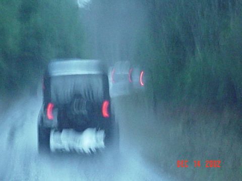 1989 Ford Bronco - A dark SUV drives in the rain, with another vehicle's taillights visible ahead.