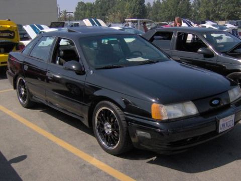 1991 Ford Taurus - Black 1991 Ford Taurus sedan with aftermarket wheels is parked in a lot with other vehicles.