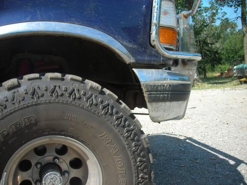 1994 Ford Bronco - Close-up of a blue Ford truck's front corner, showing its tire and bumper, with another vehicle visible in the distance.