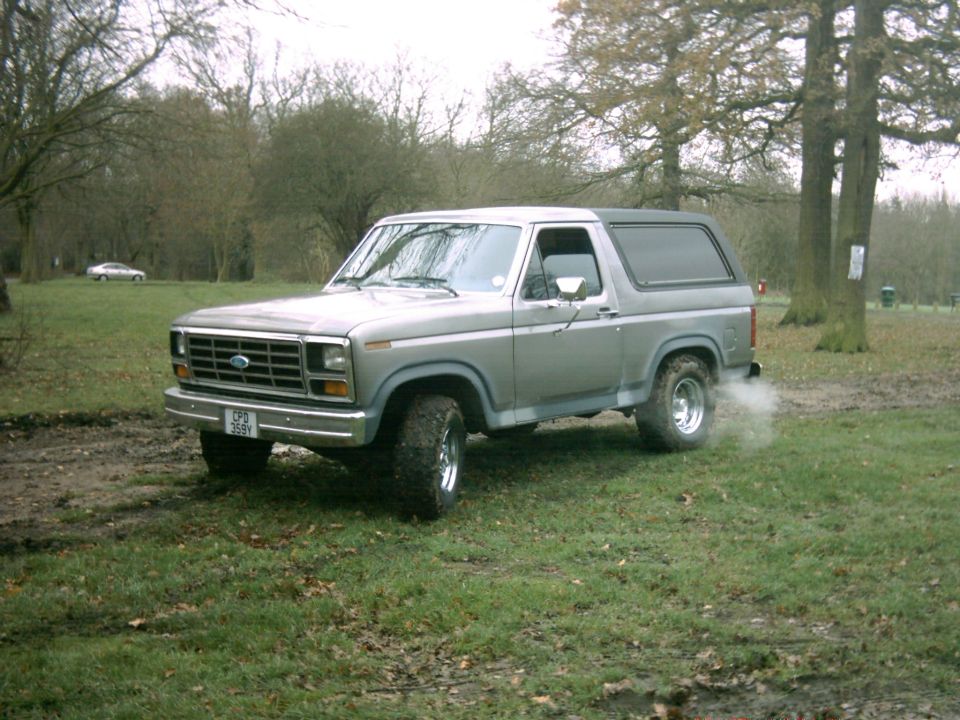 Silver Ford Bronco with aftermarket wheels and tires, seen from a 3/4 front angle in a grassy, muddy field