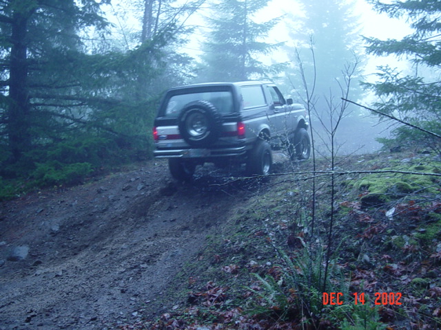 Lifted Ford SUV with rear-mounted spare descending a muddy trail in fog, pickup ahead