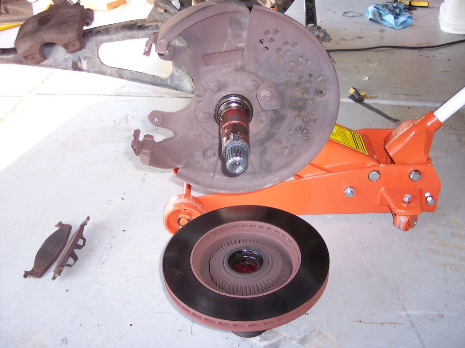 Disassembled front brake with exposed spindle and dust shield, old rotor and pads on garage floor next to orange floor jack
