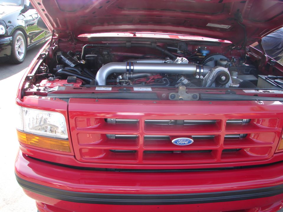 Red Ford truck with hood open, showing engine and front grille. Another black vehicle is partially visible to the left