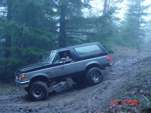 1989 Ford Bronco - A two-tone Ford Bronco navigates a wet, off-road path through a forest.