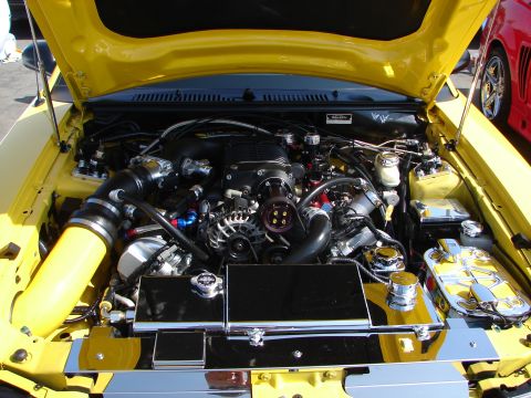 1992 Ford Taurus - The engine bay of a yellow car is open, revealing a supercharged engine and custom chrome components. Another car is visible in the backgro…
