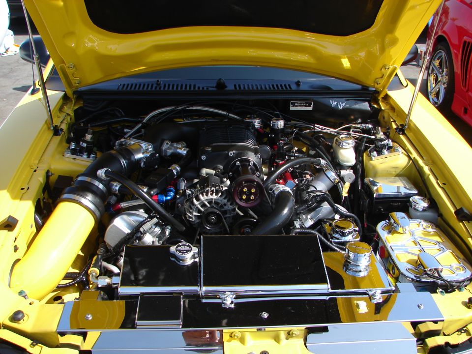 Yellow car engine bay with a supercharger, chrome parts, and a red car visible in the background