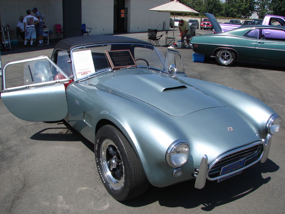 Light blue convertible with doors open, hood scoop, and chrome bumpers. Other cars and people in background