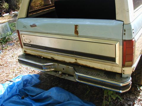 1986 Ford Bronco - The rear of a light blue SUV shows significant rust on the bumper and tailgate trim, partially covered by a blue tarp.