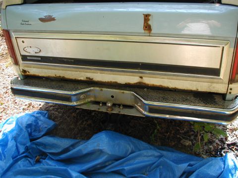 1986 Ford Bronco - The rear of a pickup truck shows significant rust around the bumper and tailgate, with a blue tarp spread out below.