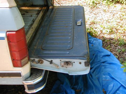 1986 Ford Bronco - The tailgate of a 1986 Ford Bronco is open, revealing its internal latching hardware and a blue tarp underneath.