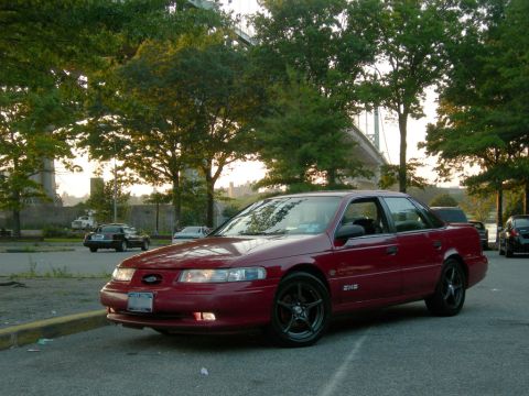 1992 Ford Taurus - A red sedan is parked in a lot near a bridge, with other vehicles in the background.