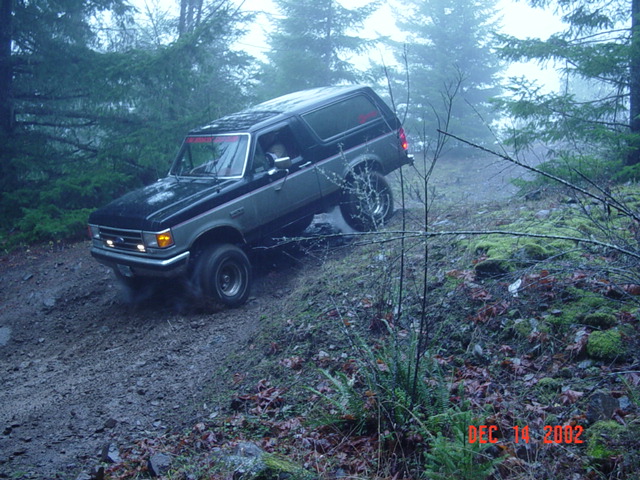 Black Ford pickup with camper shell tilted on a steep muddy embankment in foggy woods