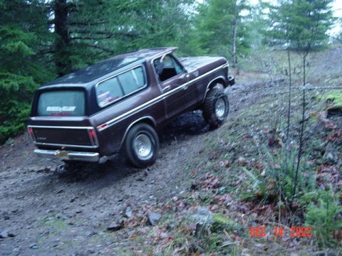 1989 Ford Bronco - A brown 1989 Ford Bronco navigates a muddy, wooded incline, its off-road tires gripping the terrain.