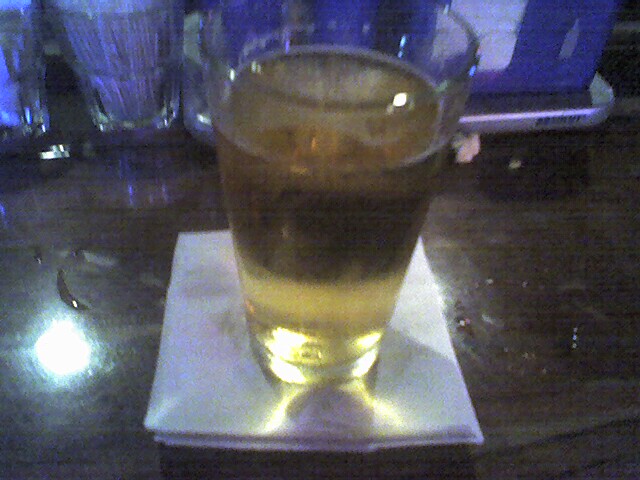 Glass of amber liquid with ice on a white napkin, with other glasses blurred in the background