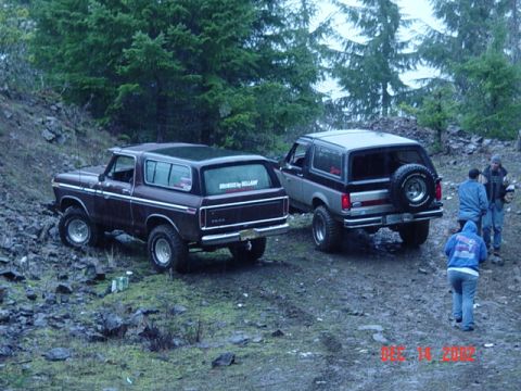 1989 Ford Bronco - Two Ford Broncos are parked on a muddy, rocky incline with trees in the background.