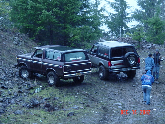 Maroon and gray lifted Ford Broncos on a wet rocky forest trail with three people standing nearby
