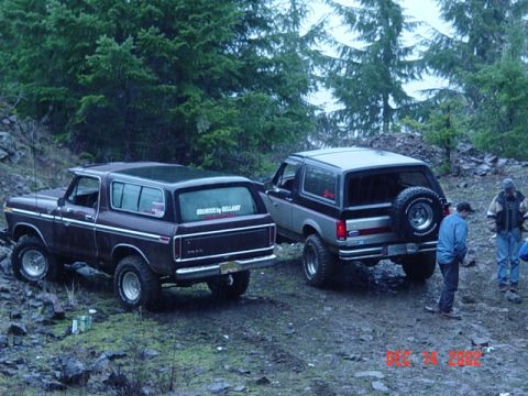 1989 Ford Bronco - Two Ford Broncos, one dark brown and one silver, are parked on a dirt trail with trees in the background.