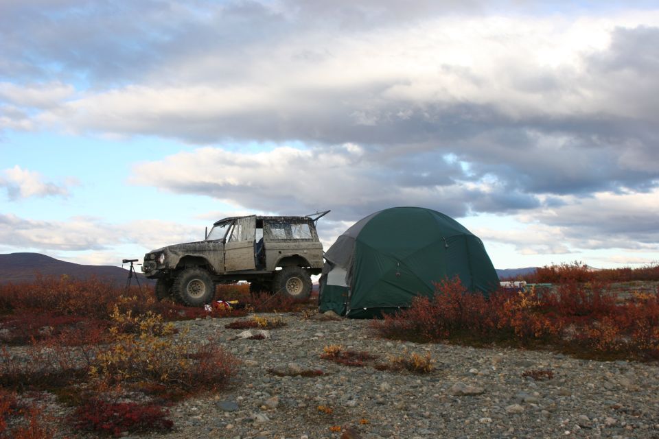 1971 Ford Bronco parked next to a green tent on a rocky, brush-covered hillside under a cloudy sky