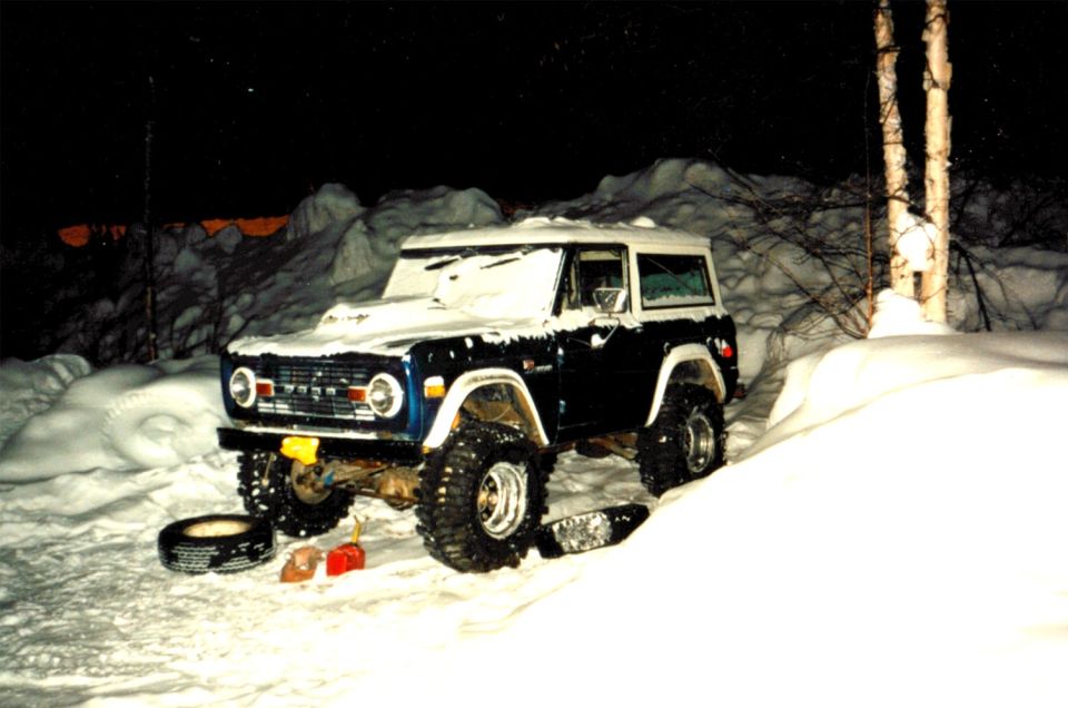 1971 Ford Bronco covered in snow, parked on a snowy incline with a spare tire and tools nearby