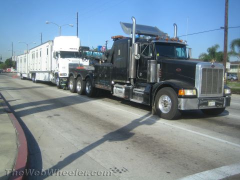 1990 Ford Bronco - black Peterbilt heavy-duty wrecker tows a semi tractor and white box trailer along a city street.