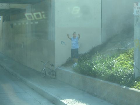 1990 Ford Bronco - View through tinted glass of a man making peace signs beside a parked bicycle along a sloped underpass embankment.