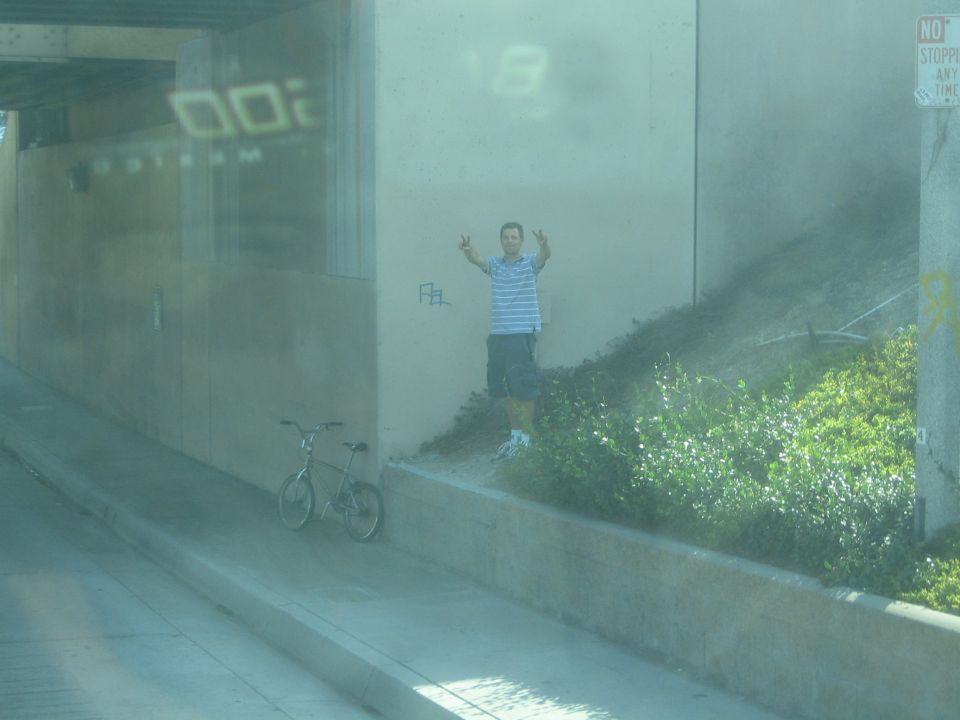 Man flashing peace signs by a bicycle under an overpass, seen through tinted glass
