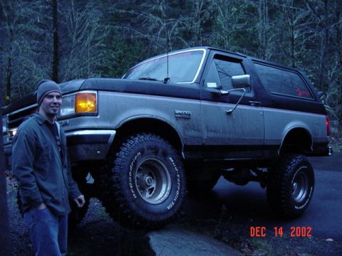 1989 Ford Bronco - Man standing next to a 1989 Ford Bronco on a dirt road in a wooded area.