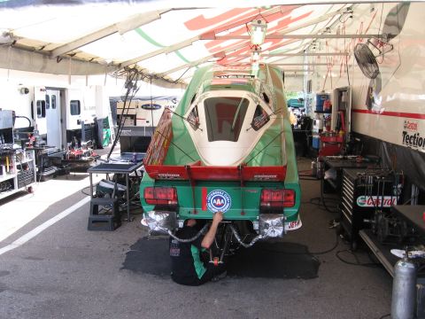 1998 GMC Sierra 1500 - A mechanic works on the rear of a green drag car in a pit stall, surrounded by equipment and support vehicles.