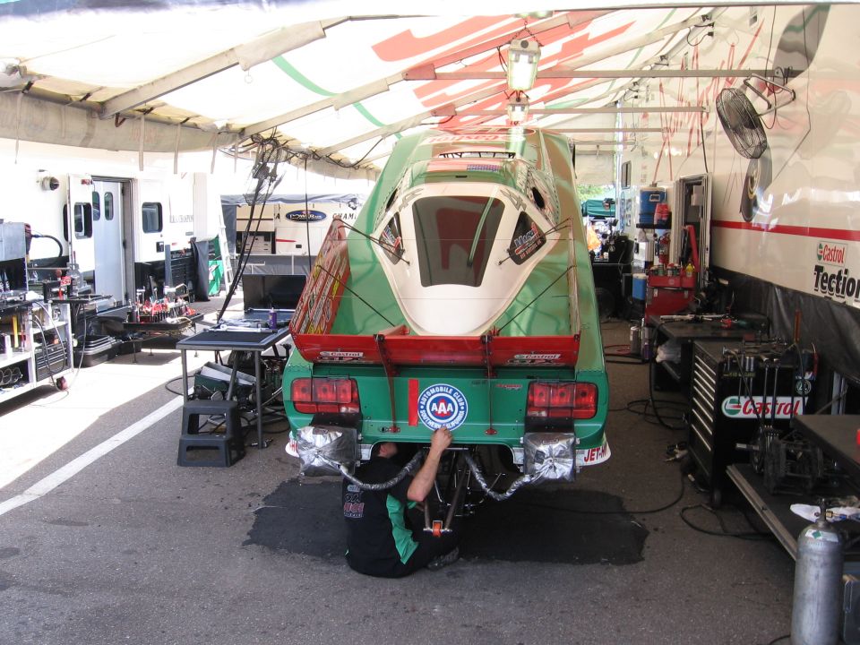 Rear view of a green drag car with a white canopy, undergoing maintenance in a pit stall