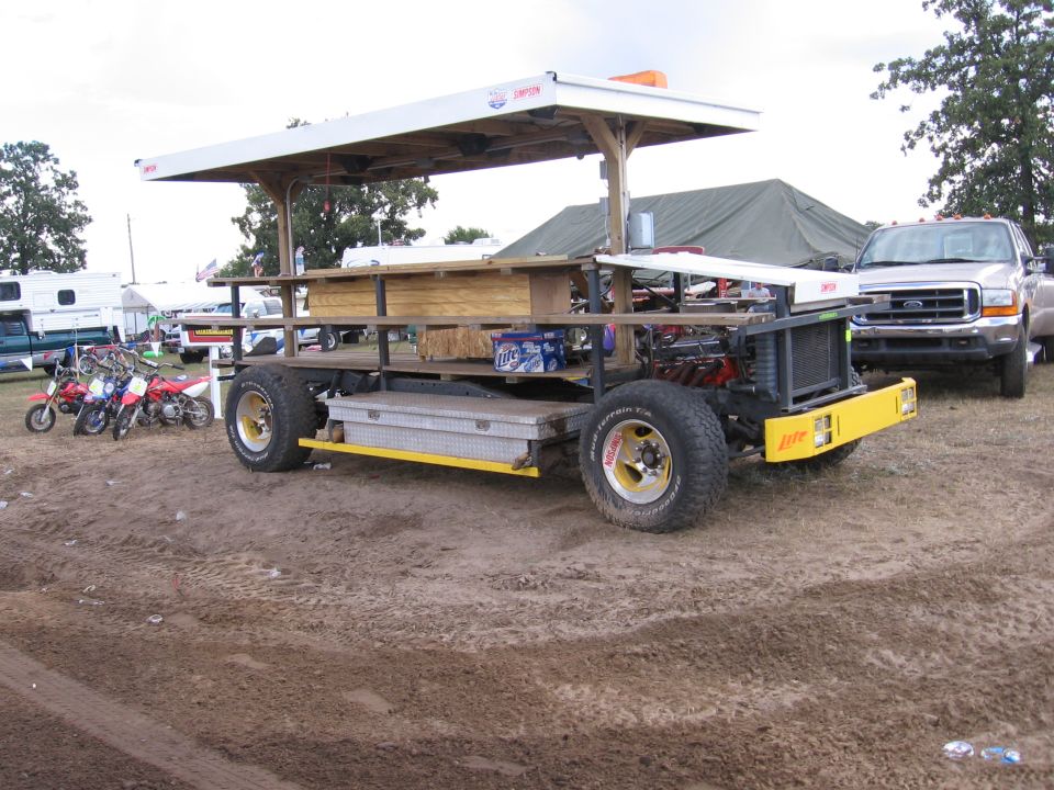 Custom vehicle with a wooden bar structure and canopy, parked next to a pickup truck and motorcycles